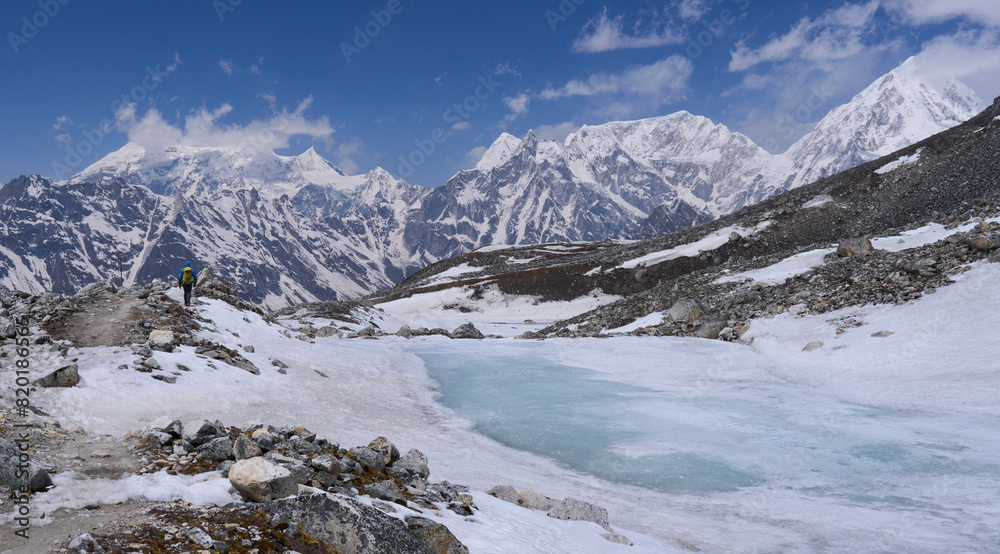 Fototapeta premium A hiker on a trail to the Tilicho Lake, Annapurna Conservation area, Nepal. A small lake covered by ice. A trail covered by snow and ice. Frozen lake. Mountain range in the background.