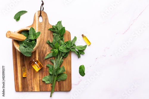 Wooden mortar with fresh green mint leaves and bottles with essential oils on the wooden board on white marble table top view, copy space for your design. Herbal and aromatheraphy concept. 