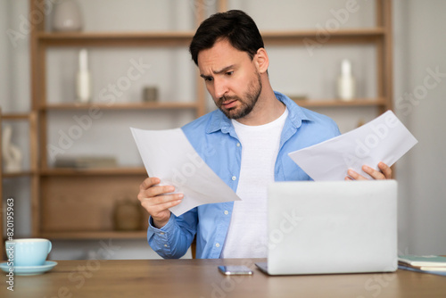 A man is sitting at a desk at home office, looking with a perplexed expression at some documents in his hands. A laptop is open in front of him, suggesting he is cross-referencing information