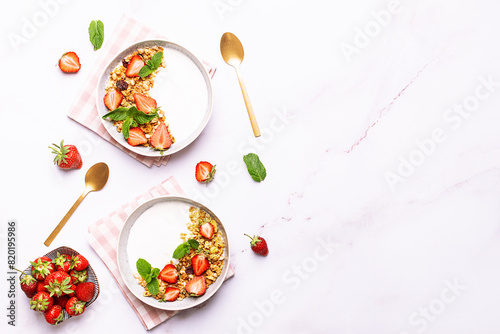 Fresh Greek Yogurt with granola and strawberries decorated with aromatic mint leaves on white marble table top view, copy space for your design. Healthy breakfast concept.