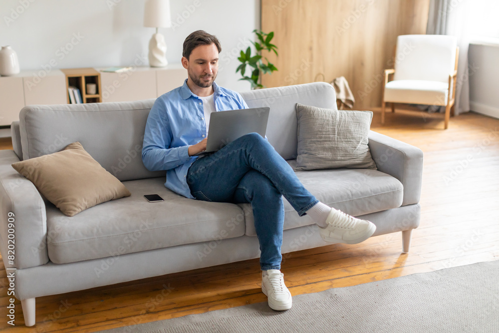 A man is seated on a couch, focused on his laptop screen. He appears engaged in his work or activities online, with a serious expression. The room is simple, with no other visible people A man is seated on a couch, focused on his laptop screen. He appears engaged in his work or activities online, with a serious expression. The room is simple, with no other visible people