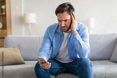 A man appears troubled or exasperated, sitting with a slouched posture on a grey couch. He cradles his head in one hand, with a smartphone held in the other. His expression conveys distress or concern photo