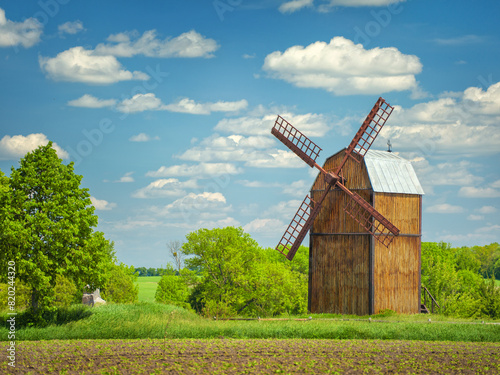 view to wooden windmill in farm field under blue sky with white clouds in spring day