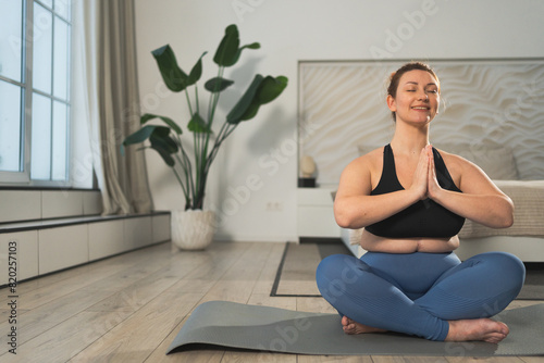 Yoga mindfulness meditation. Woman practicing yoga in living room at home. Plus size female sitting in lotus pose meditating smiling relaxing indoor. Girl doing breathing practice. Yoga at home