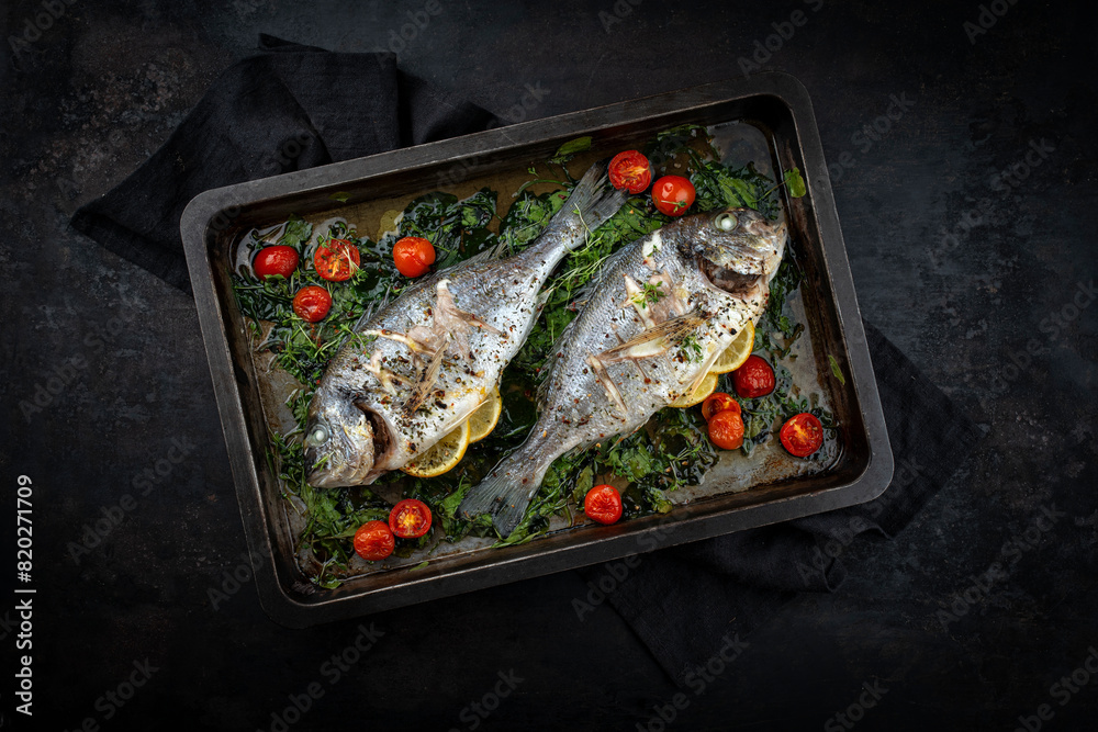 Two fresh steamed gilthead seabreams with tomatoes and lettuce served as top view in a rustic backing sheet Two fresh steamed gilthead seabreams with tomatoes and lettuce served as top view in a rustic backing sheet