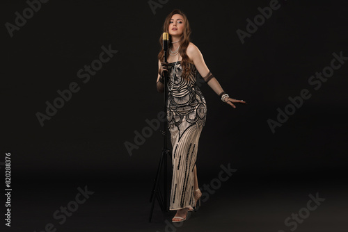 Beautiful young woman in stylish dress with microphone singing on black background