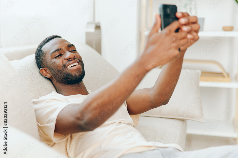 Smiling African American Man Using Black Smartphone on Modern White Sofa at Home The man is casually dressed in a white tshirt, lying down comfortably while typing a message on his mobile phone With a