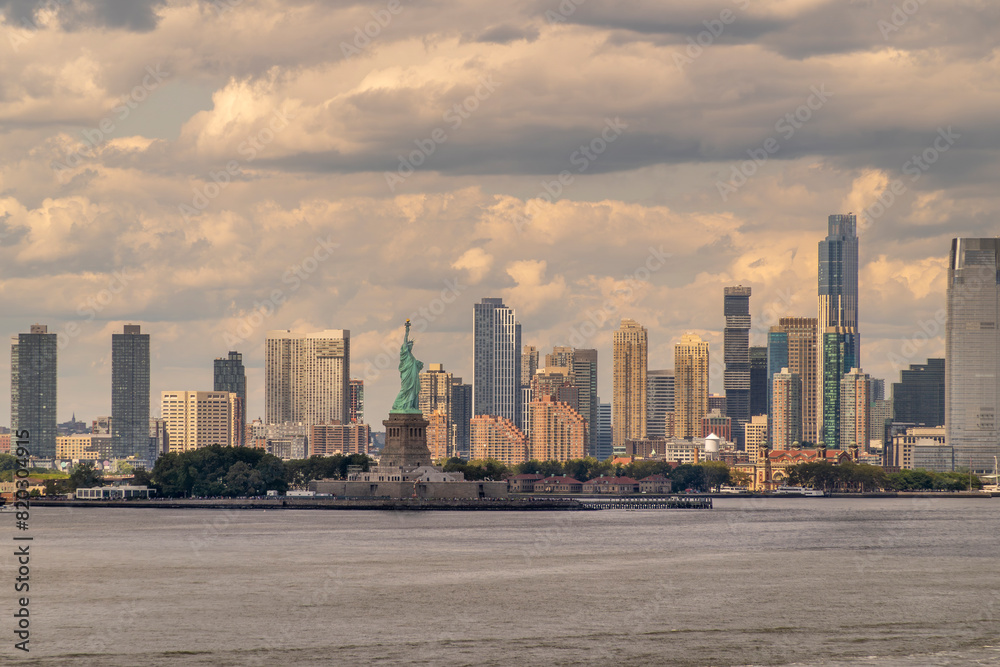New York, NY, USA - August 1, 2023: Statue of Liberty and its green island under blue cloudscape. Skyline of Jersey City with wall of tall buildings New York, NY, USA - August 1, 2023: Statue of Liberty and its green island under blue cloudscape. Skyline of Jersey City with wall of tall buildings