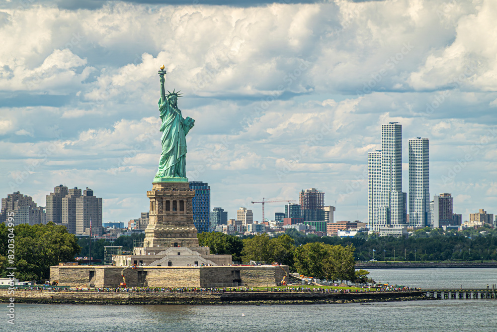 Obraz premium New York, NY, USA - August 1, 2023: Jouned Squared tower of Jersey city is seen to the right of Statue of Liberty on its island under blue cloudscape. Plenty of visitors and more building in back