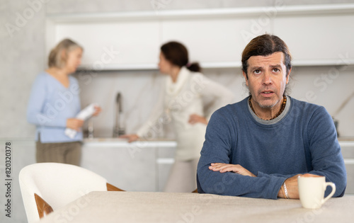 Thinking man sits in kitchen and in background adult daughter quarrels with elderly mother