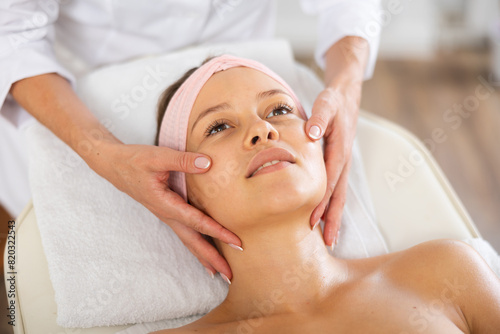 Female client is resting during massage face skin. Female physiotherapist performs restorative stroking during spa care.