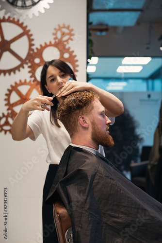 Red-haired bearded man sitting in barbershop and female barber making hairdo to guest
