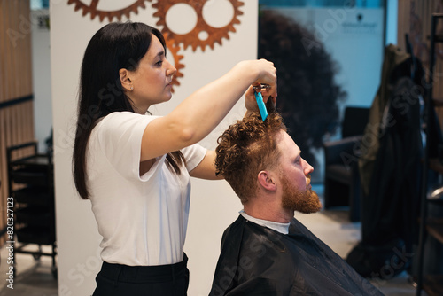 Red-haired bearded man sitting in barbershop and female barber making hairdo to guest