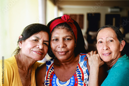 Happy senior people looking at camera at home. Multiracial older friends hug each other. Focus on black woman face