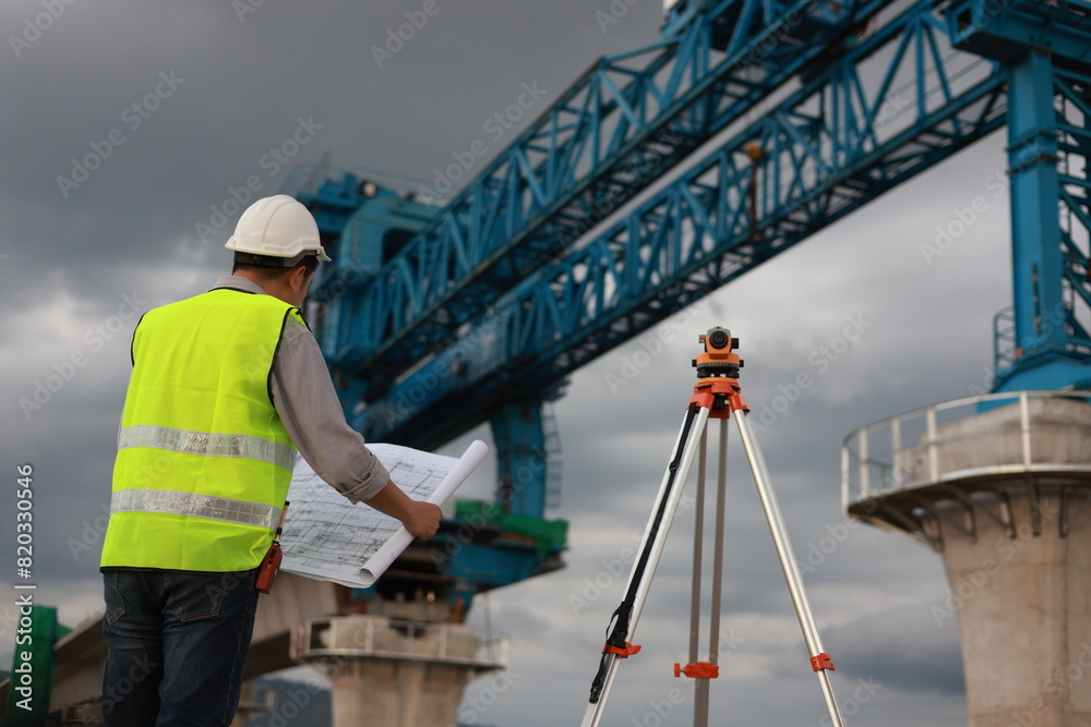 Senior Construction Engineer is Holding Paper Plans, Blueprint, and Talking with Coworkers Using a survey camera at the construction site. Concrete cement pillar construction of Skytrain a mass rail t Senior Construction Engineer is Holding Paper Plans, Blueprint, and Talking with Coworkers Using a survey camera at the construction site. Concrete cement pillar construction of Skytrain a mass rail t