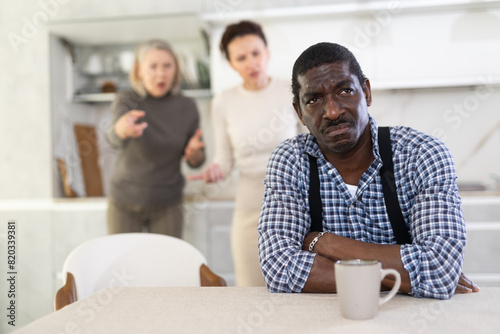 Offended middle-aged man sitting in the kitchen and two women brawling to him angrily standing behind photo