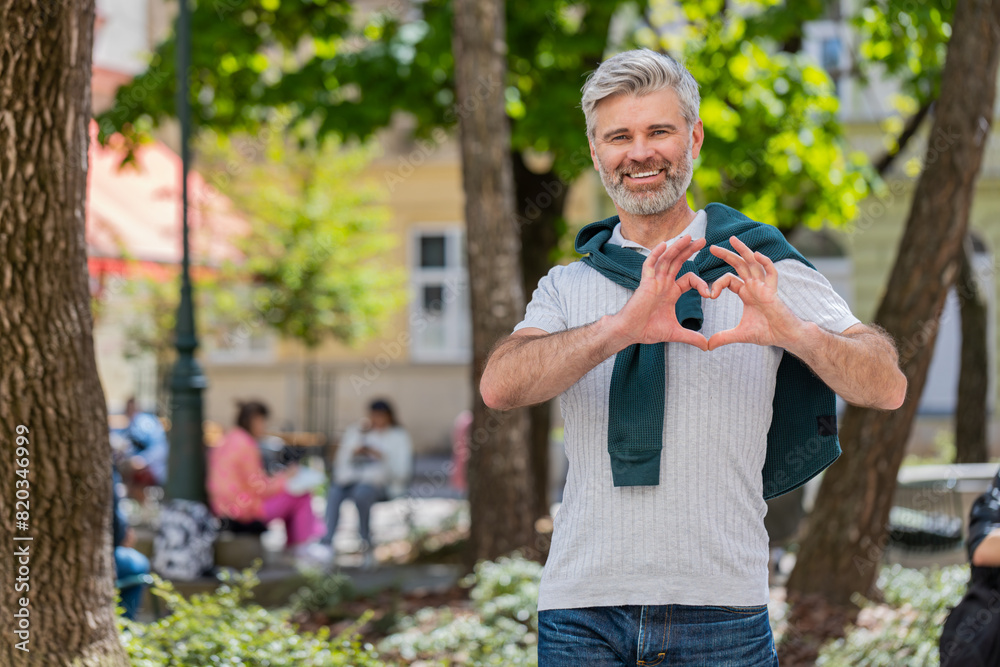 Fototapeta premium I love you. Caucasian middle-aged man makes symbol of love showing heart sign to camera, express sincere romantic positive feelings. Charity, gratitude, donation. Mature guy on urban city street park.