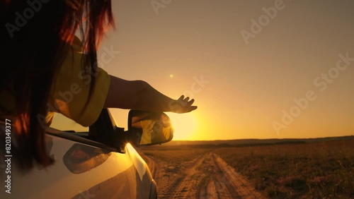 Beautiful girl with sits in front seat of car, her hand out window, catching glare of setting sun. Free young woman travels by car, extends her hand to sun from car window. Vacation. Woman driver sun photo