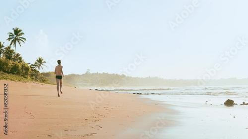 Brazilian man wearing swimwear running on the sand of a paradisiacal beach, with coconut trees and sea waves in the background photo