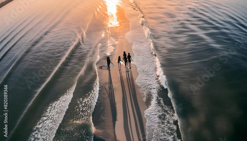 Aerial view of three surfers walking along the beach at sunset, capturing their shadows in the water	