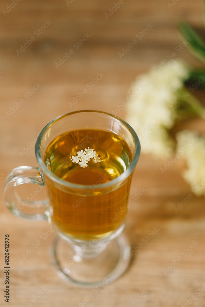 A clear glass mug filled with elderflower tea is placed on a rustic wooden table. Fresh elderflowers and a wooden cutting board are nearby, creating a cozy and natural atmosphere. A clear glass mug filled with elderflower tea is placed on a rustic wooden table. Fresh elderflowers and a wooden cutting board are nearby, creating a cozy and natural atmosphere.
