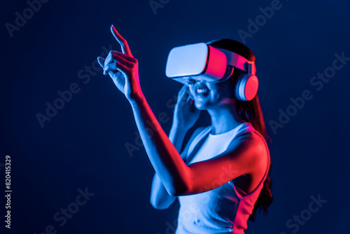 Smart female stand with surrounded by cyberpunk neon light wear VR headset connecting metaverse, futuristic cyberspace community technology. Woman using finger pointing virtual object. Hallucination. © Summit Art Creations