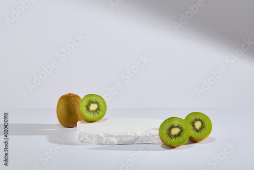 Against white background, a stone podium is decorated with some halves of kiwi fruit. Empty space for product presentation. Abstract composition in minimal design
