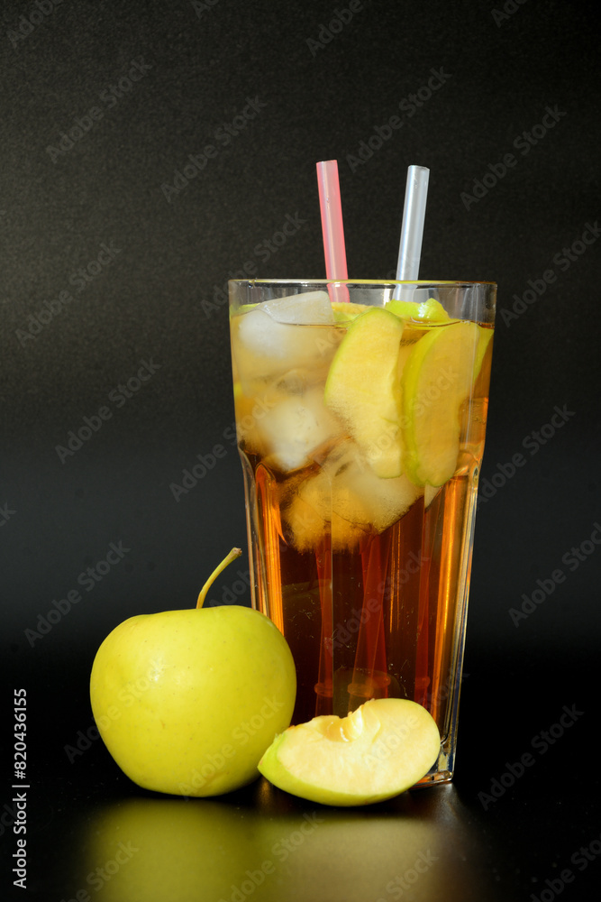 Fototapeta premium A faceted tall glass of apple juice with ice on a black background, next to ripe slices of a green apple.