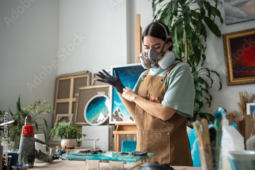 Creative female artist wearing respirator, latex gloves works with epoxy resin, crafting pieces on canvas in art studio. Manufacturing process of products made of liquid decorating material  photo