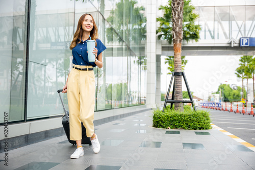 A young Asian woman with a sense of wanderlust holds a refreshing beverage in one hand and her suitcase in the other. Ready for travel, she sets off on her next adventure.