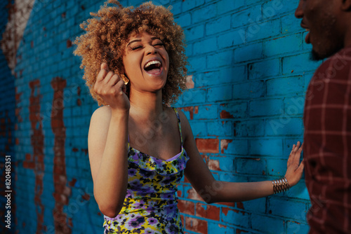 A joyful woman with curly hair laughing and dancing in front of a blue brick wall, interacting playfully with a man partially visible.