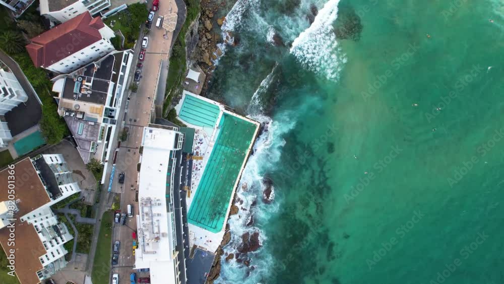 A birds eye drone view looking down on the Bondi Icebergs Club on Bondi Beach in Sydney, New South Wales. A day club and swimming pool where you can look out at the blue ocean and waves crashing. A birds eye drone view looking down on the Bondi Icebergs Club on Bondi Beach in Sydney, New South Wales. A day club and swimming pool where you can look out at the blue ocean and waves crashing.