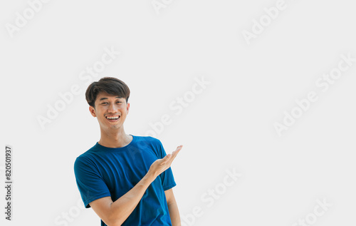 A young Asian man in his 20s wearing a blue t-shirt making a welcome gesture or presenting isolated on a gray background. Space for advertisements or welcome messages. Happy man. Positive person.