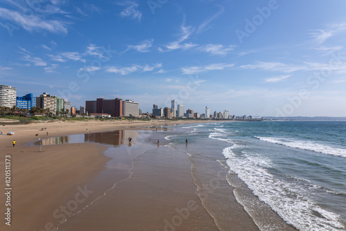 A perspective view of the beachfront with waves, exposed beach sand at low tide, city buildings and a blue sky with some clouds in the tourist city of Durban in KwaZulu Natal in South Africa.