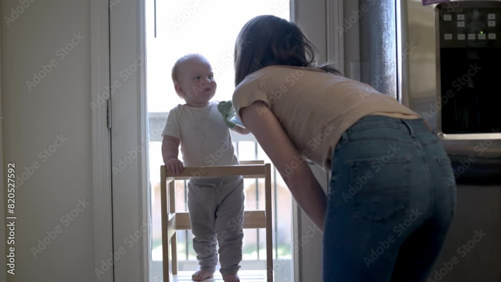 This tender scene captures a toddler in a learning tower offering a sippy cup to his mother, highlighting a moment of sharing and connection in a sunlit kitchen, symbolizing early social development. This tender scene captures a toddler in a learning tower offering a sippy cup to his mother, highlighting a moment of sharing and connection in a sunlit kitchen, symbolizing early social development.