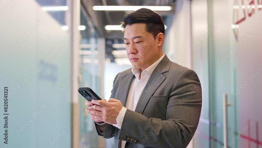 Side view portrait of confident japanese male browsing personal smartphone in office environment. Serious young businessman holding modern gadget indoors. Concept of technology and occupation. Side view portrait of confident japanese male browsing personal smartphone in office environment. Serious young businessman holding modern gadget indoors. Concept of technology and occupation.
