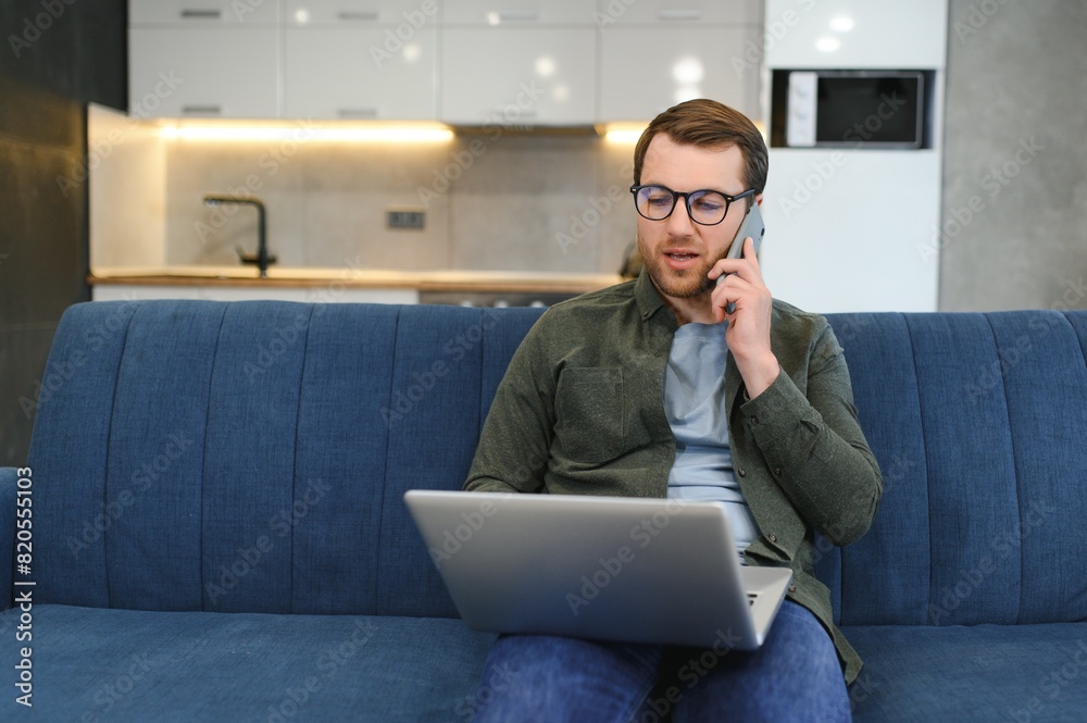 Cheerful busy young caucasian male manager sits on sofa with computer, calls by phone, talks with client, looks at free space in white room interior. Business, work remotely at home, ad and offer. Cheerful busy young caucasian male manager sits on sofa with computer, calls by phone, talks with client, looks at free space in white room interior. Business, work remotely at home, ad and offer.