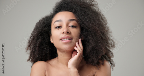 Beauty and healthcare concept - beautiful African American woman with curly afro hairstyle and clean, healthy skin touches her cheek and face with her hand, posing and looking at the camera