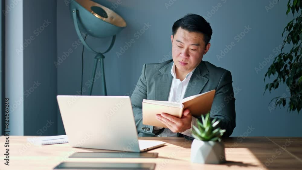Confident chinese male having online conversation on modern laptop and reading information from personal notebook indoors. Competent young businessman conducting video call in well-lit cabinet.