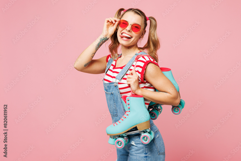 Side view young happy woman she wear red t-shirt denim overalls casual clothes glasses hold blue rollers show tongue isolated on plain pastel pink background. Summer sport lifestyle leisure concept.