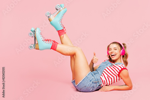 Foto Full body young woman wears red t-shirt overalls casual clothes blue rollers rollerblading lay down raise up legs listen to music in headphones isolated on plain pink background