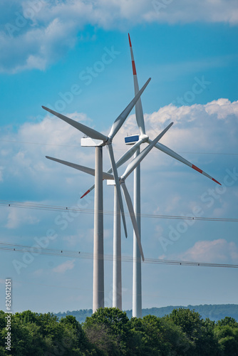 Drei, perspektivisch eng zusammenstehende Windräder zur Erzeugung nachhaltigen Stroms mit Überlandleitung, Baumkronen und leicht bewölktem, dunstigen Himmel