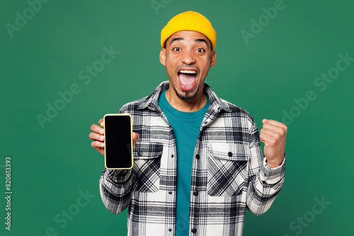 Young man of African American ethnicity in shirt t-shirt yellow hat hold use mobile cell phone with blank screen workspace area do winner gesture isolated on plain green background Lifestyle concept