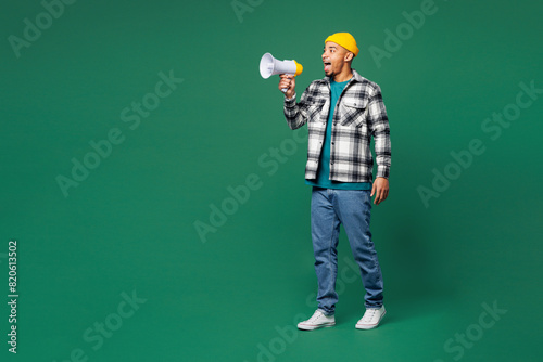 Young man of African American ethnicity wear shirt blue t-shirt yellow hat hold in hand megaphone scream announces discounts sale Hurry up isolated on plain green background studio. Lifestyle concept.