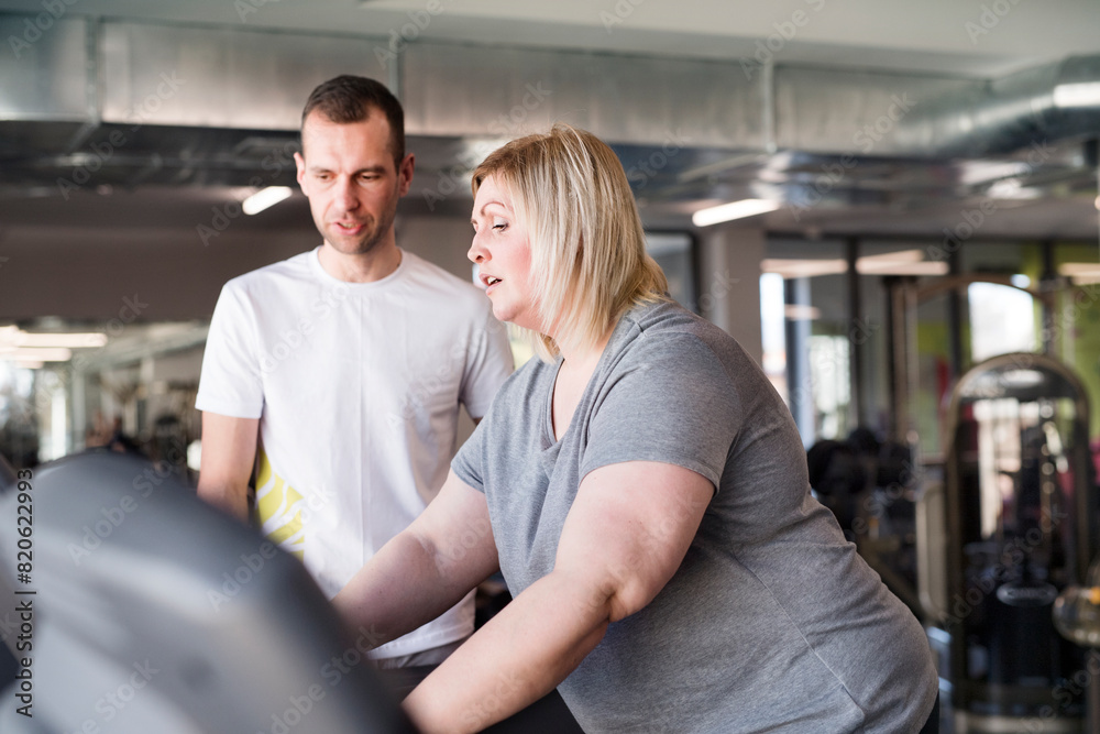 Overweight woman exercising on treadmill in gym. Personal trainer helping her. Overweight woman exercising on treadmill in gym. Personal trainer helping her.