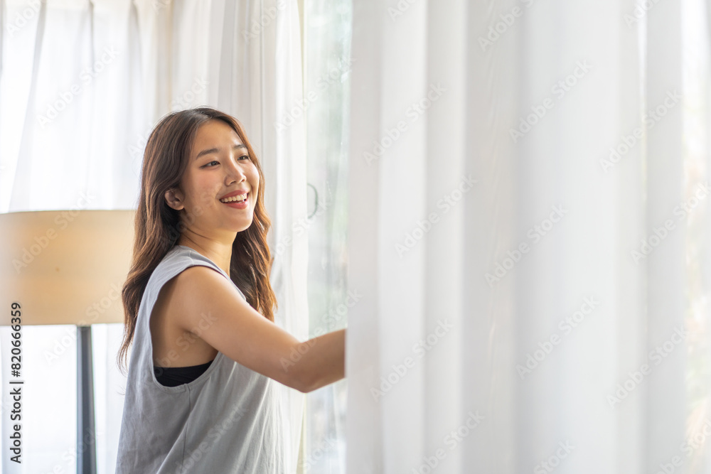 Happy young beautiful pretty asian woman waking up and opening window curtains.Girl feeling comfortable and relaxed breathing fresh air in morning in bedroom and copy space banner at home Happy young beautiful pretty asian woman waking up and opening window curtains.Girl feeling comfortable and relaxed breathing fresh air in morning in bedroom and copy space banner at home