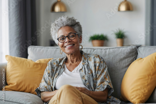 A happy 60-year-old a gray-haired African American woman is relaxing on a light gray sofa against the blurred background of a bright living room