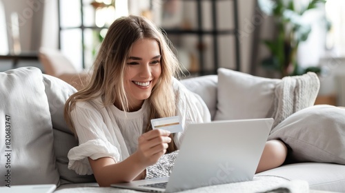 A smiling woman is shopping online at home using a credit card and laptop on the couch. This modern lifestyle is characterized by convenience, internet transactions, and secure online payments