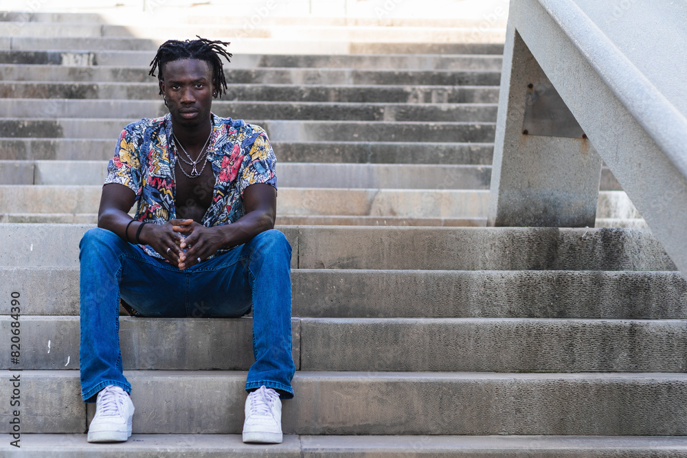 Low angle full body of serious African American male in colorful shirt and jeans with sneakers sitting on stone steps and looking at camera on urban street Low angle full body of serious African American male in colorful shirt and jeans with sneakers sitting on stone steps and looking at camera on urban street
