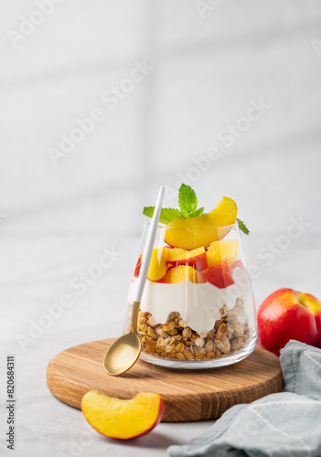 Greek yogurt parfait with peach and muesli in a glass on a wooden board on a light background with fresh fruits, napkin and shadow.